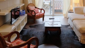 Un salon comprenant un canapé, deux fauteuils, une table basse et un téléviseur posé sur un meuble. On y trouve également des étagères remplies de livres et d’objets décoratifs, ainsi qu’une fenêtre avec des rideaux.
