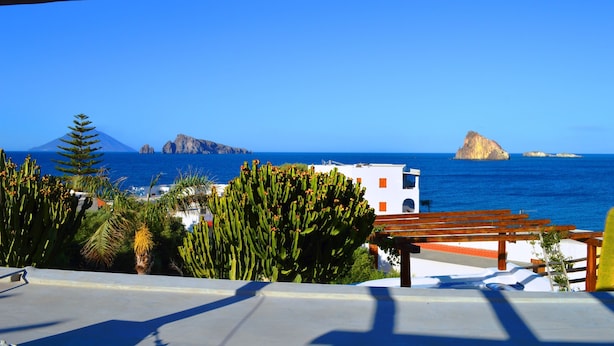 A coastal view with a wooden pergola, cacti, and a white building with orange accents. - Casa Ambra (Lipari)