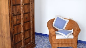 A wicker chair with two pillows against a white wall, a wooden door, and blue patterned floor tiles. - Casa Ambra (Lipari)