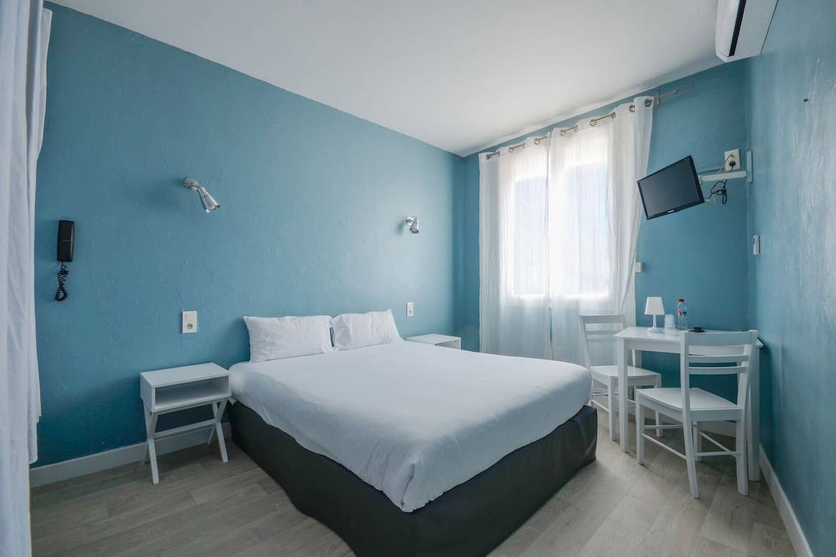A bedroom with a bed, bedside tables, a desk with chairs, a TV, and a phone on the wall.