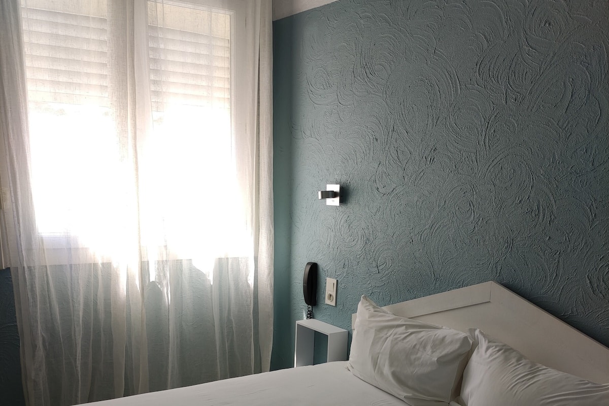 A bedroom with a bed, white curtains, a bedside table with a phone, and a textured wall.