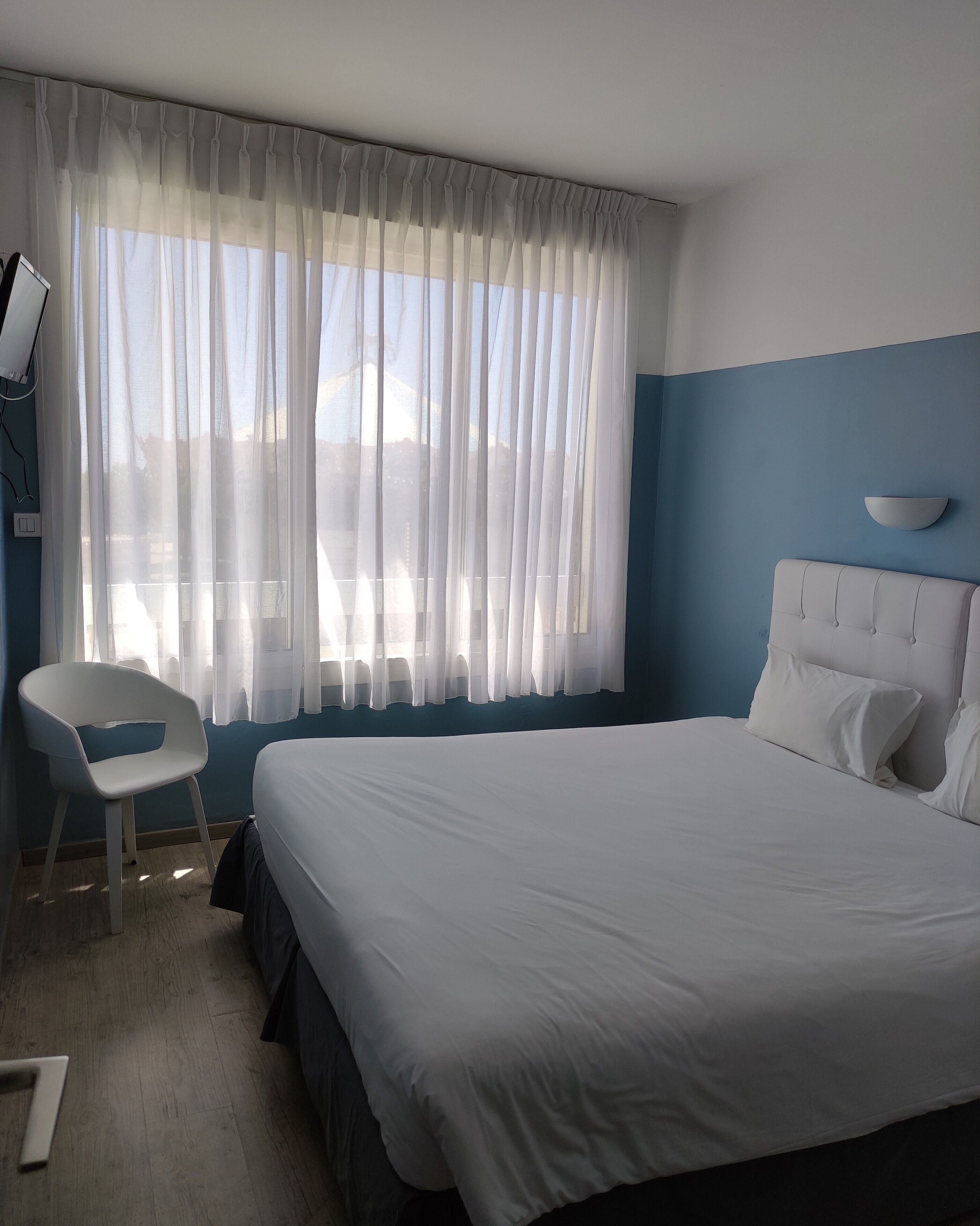 A hotel room with a large bed, a chair, a bedside table, and a window with sheer curtains.