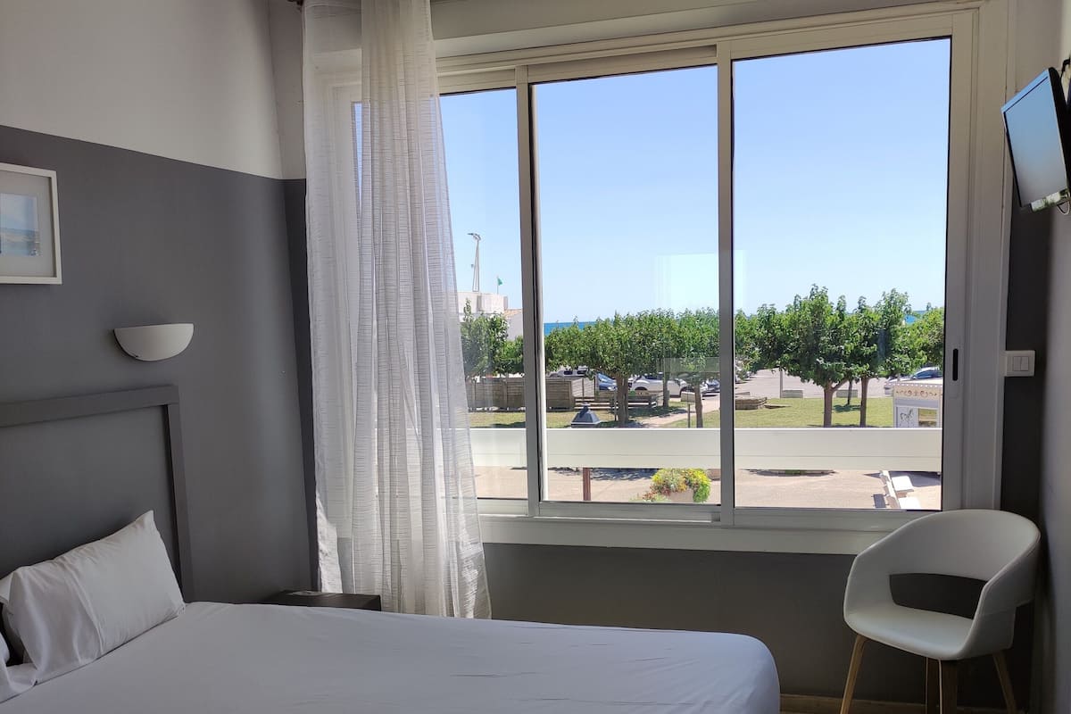 A hotel room with a large bed, a chair, a window with a view of a parking lot and trees, and a wall-mounted TV.
