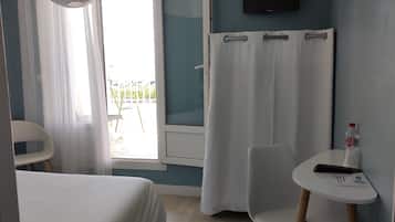 Habitación de hotel con cama, una silla, una mesita con una botella y un control remoto, un televisor en la pared y una ventana con cortinas.