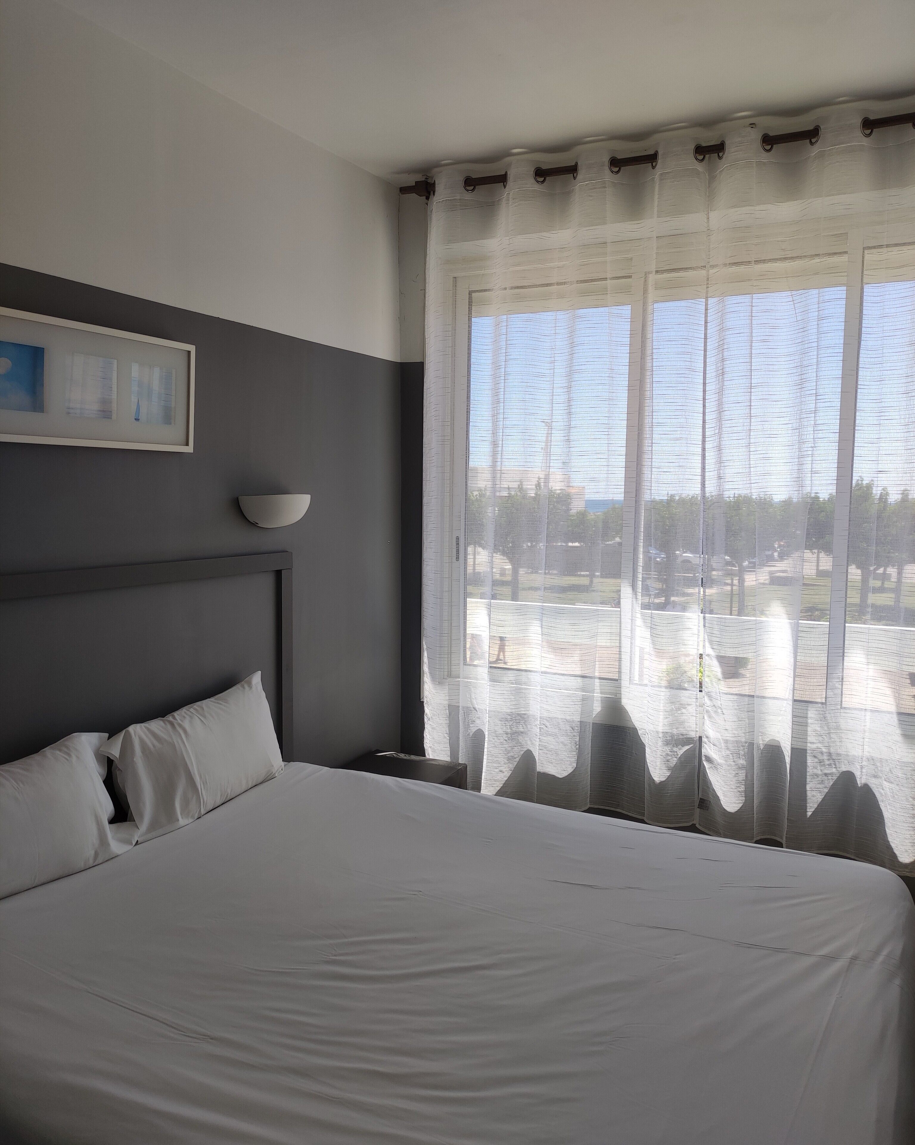 A hotel room with a large bed, a bedside table, a wall-mounted lamp, and a window with sheer curtains.