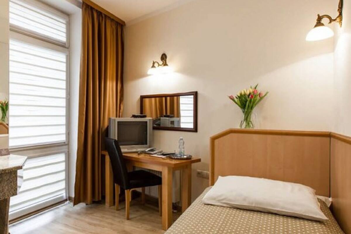 Una habitación de hotel con cama, escritorio con computadora, silla, ventana con cortinas y lámpara fijada a la pared.