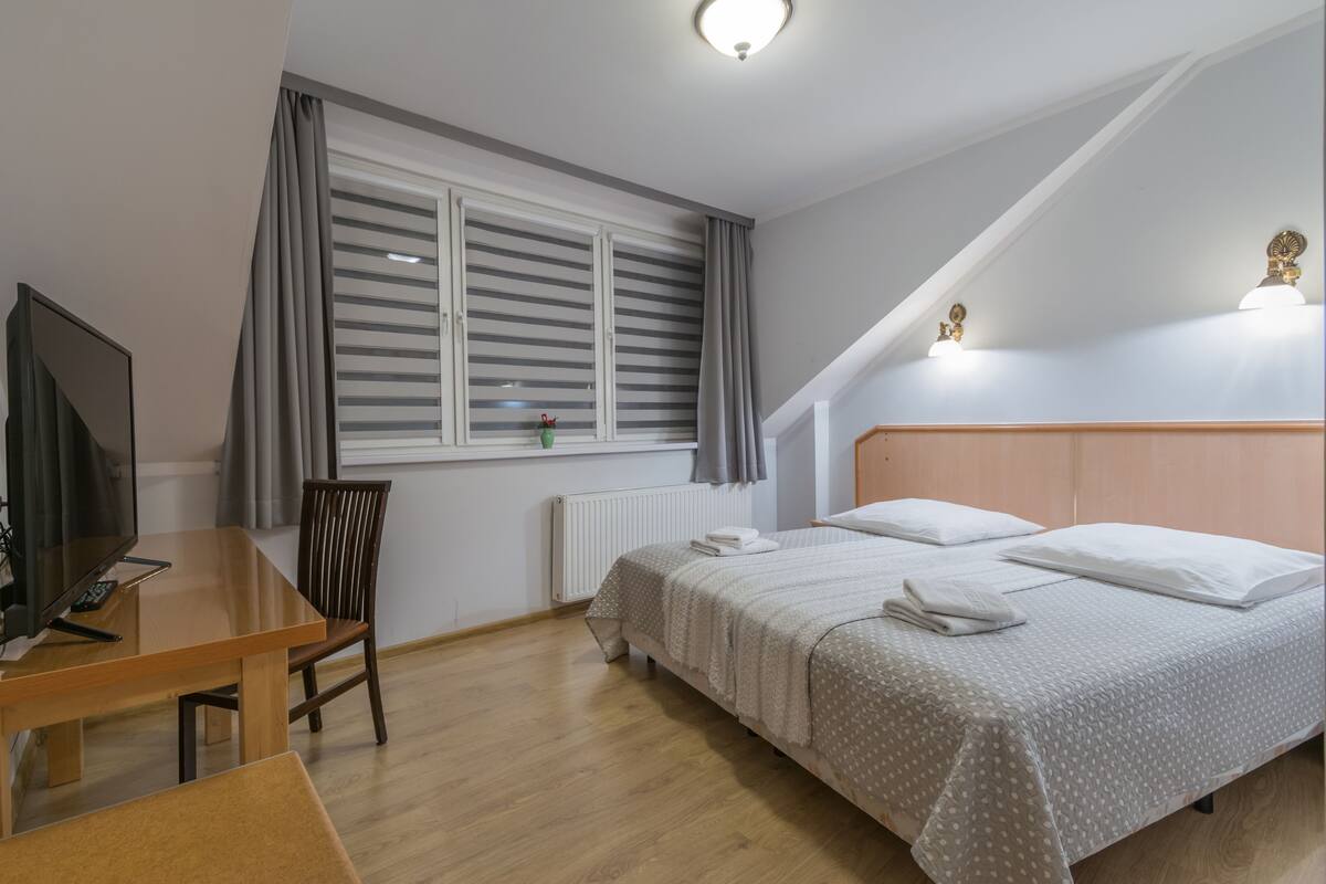 Un dormitorio con cama, escritorio, silla, televisor y ventana con cortinas.