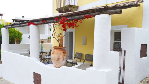 A white-walled building with a yellow facade, a large terracotta pot, and a wooden pergola with red flowers.
