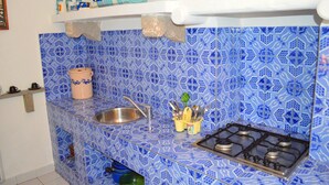 A kitchen with blue patterned tiles, a stainless steel sink, a gas stove, and a shelf with dishes and utensils.