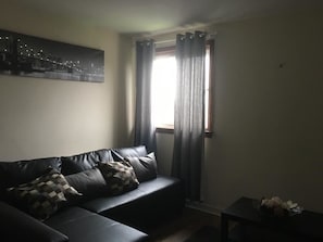 A living room with a black leather sofa, a small table with decorative items, and a window with curtains. - Majesty Tweed Apartment (Dundee)