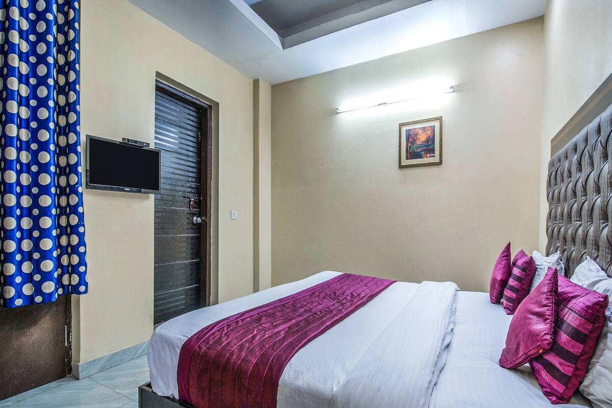 A hotel room with a bed, a television, a door, and a painting on the wall.