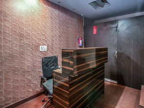 A reception area with a wooden reception desk, a black office chair, a fire extinguisher, a door with a CCTV sign, and a wall with a textured pattern. - OYO 2807 Hotel Crosswinds Residency (Noida)