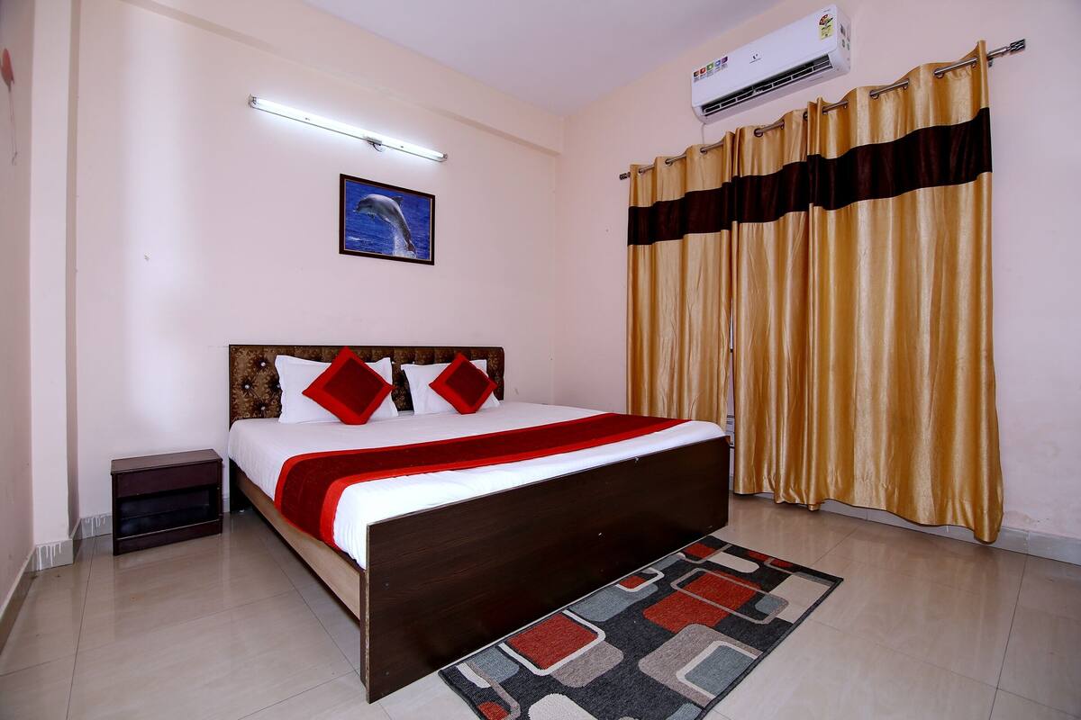 A bedroom with a bed, a nightstand, a wall-mounted air conditioner, a ceiling light, a framed picture, and a curtain.