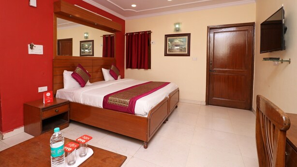 A hotel room with a wooden bed, a nightstand, a desk with a chair, a television mounted on the wall, and a door. - OYO 10661 DSR Classic (Noida)