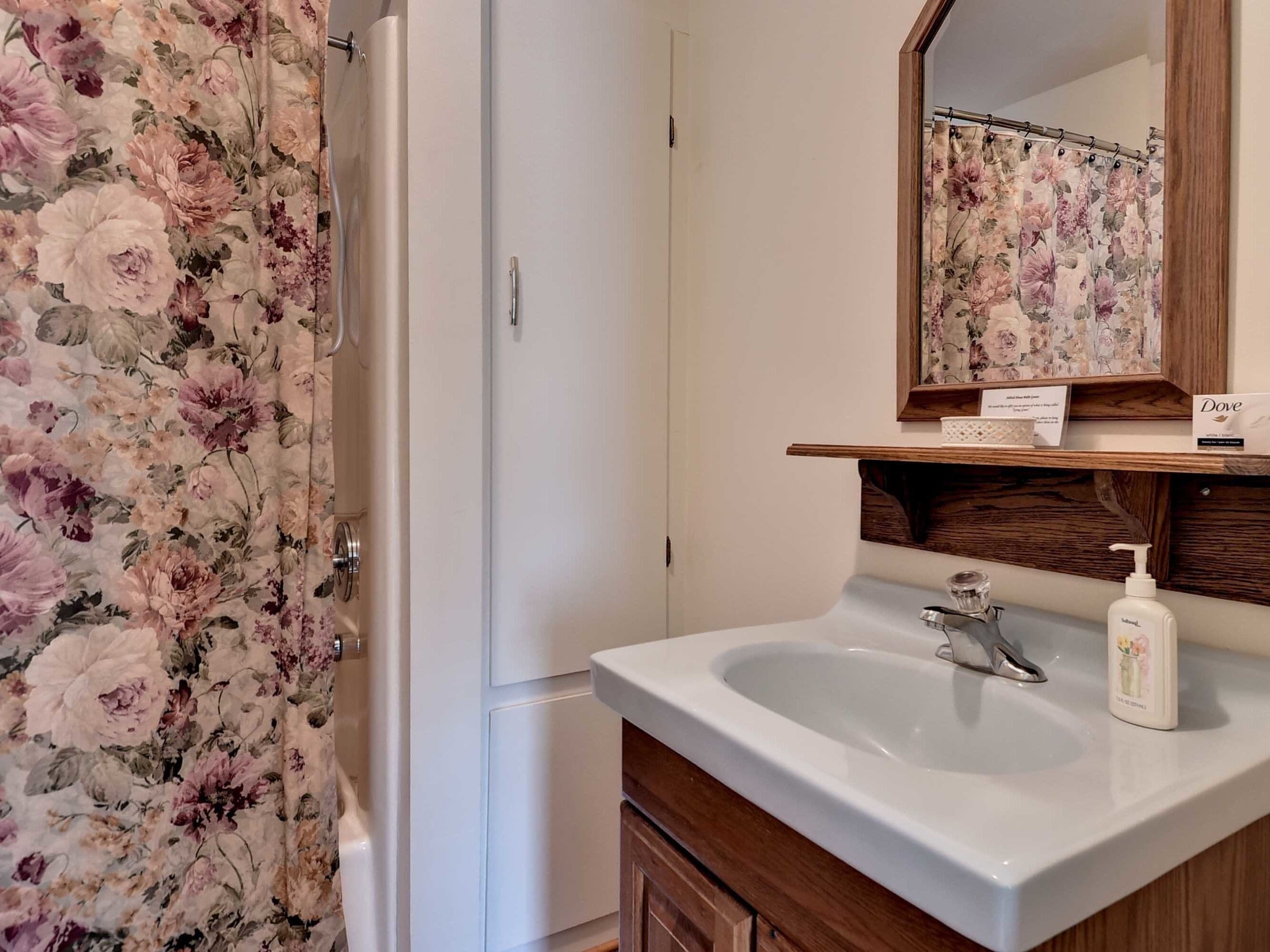 Un baño con cortina de ducha estampada con motivos florales, un lavamanos blanco con dispensador de jabón y un mueble de tocador de madera.