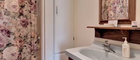 Un baño con cortina de ducha estampada con motivos florales, un lavamanos blanco con dispensador de jabón y un mueble de tocador de madera.
