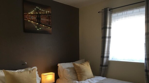 A bedroom with two beds, a nightstand with a lamp, a painting on the wall, and a window with curtains. - Majesty Weavers Loan (Dundee)