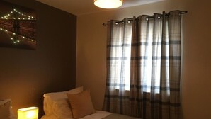 A bedroom with a bed, a nightstand with a lamp, plaid curtains, and a wall-mounted light fixture. - Majesty Weavers Loan (Dundee)