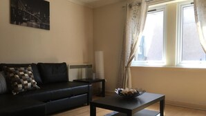 A living room with a black sofa, a small table with a bowl, a lamp, and a window with curtains. - Majesty Weavers Loan (Dundee)