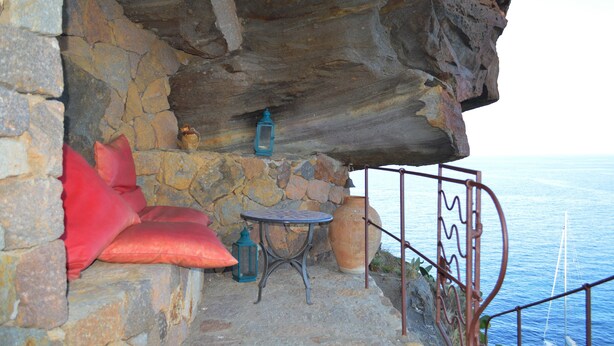 A stone-walled room with a small table and two lanterns, a red cushion, and a view of the sea. - Appartamento Zeus (Lipari)