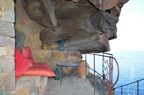 Une pièce aux murs de pierre, avec une petite table, deux lanternes, un coussin rouge et une vue sur la mer.