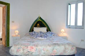 A bed with a floral bedspread, a green and yellow headboard, two bedside lamps, and a window with white frames. - Appartamento Zeus (Lipari)