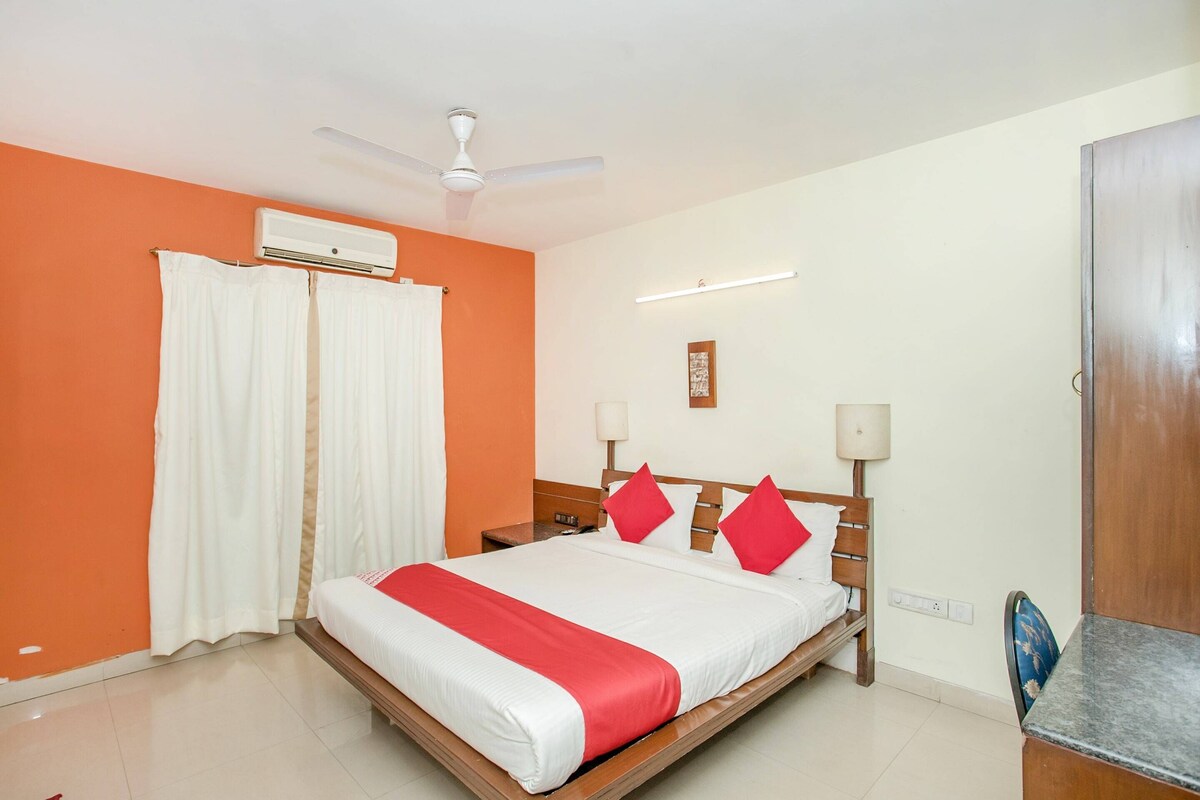 A hotel room with an orange wall, a bed with white and red bedding, a ceiling fan, an air conditioning unit, two bedside lamps, and a wooden nightstand.