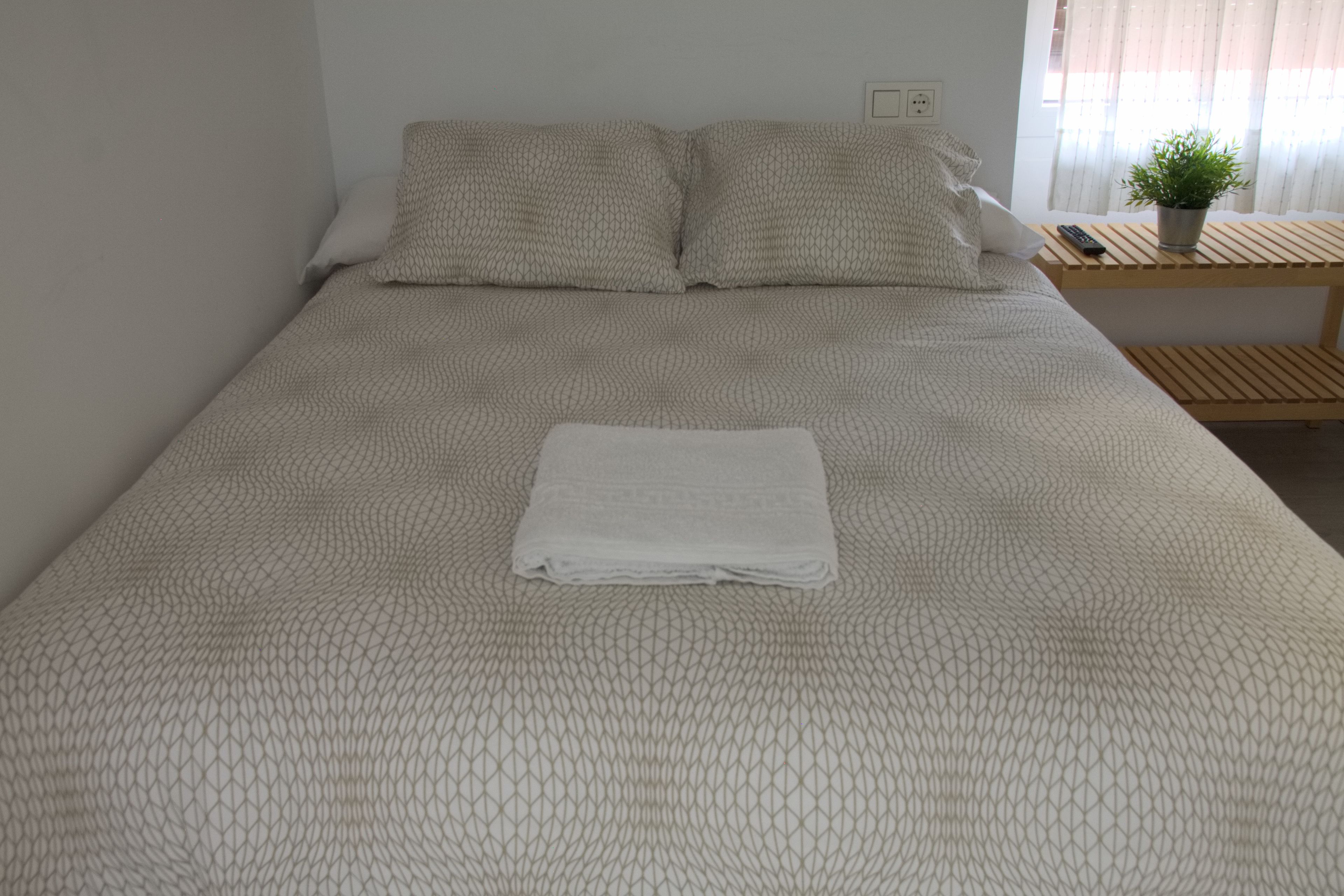 Een net opgemaakt bed met een beddengoed met een patroon, een opgevouwen witte handdoek en een nachtkastje met een plant en een afstandsbediening.