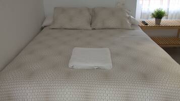 Een net opgemaakt bed met een beddengoed met een patroon, een opgevouwen witte handdoek en een nachtkastje met een plant en een afstandsbediening.
