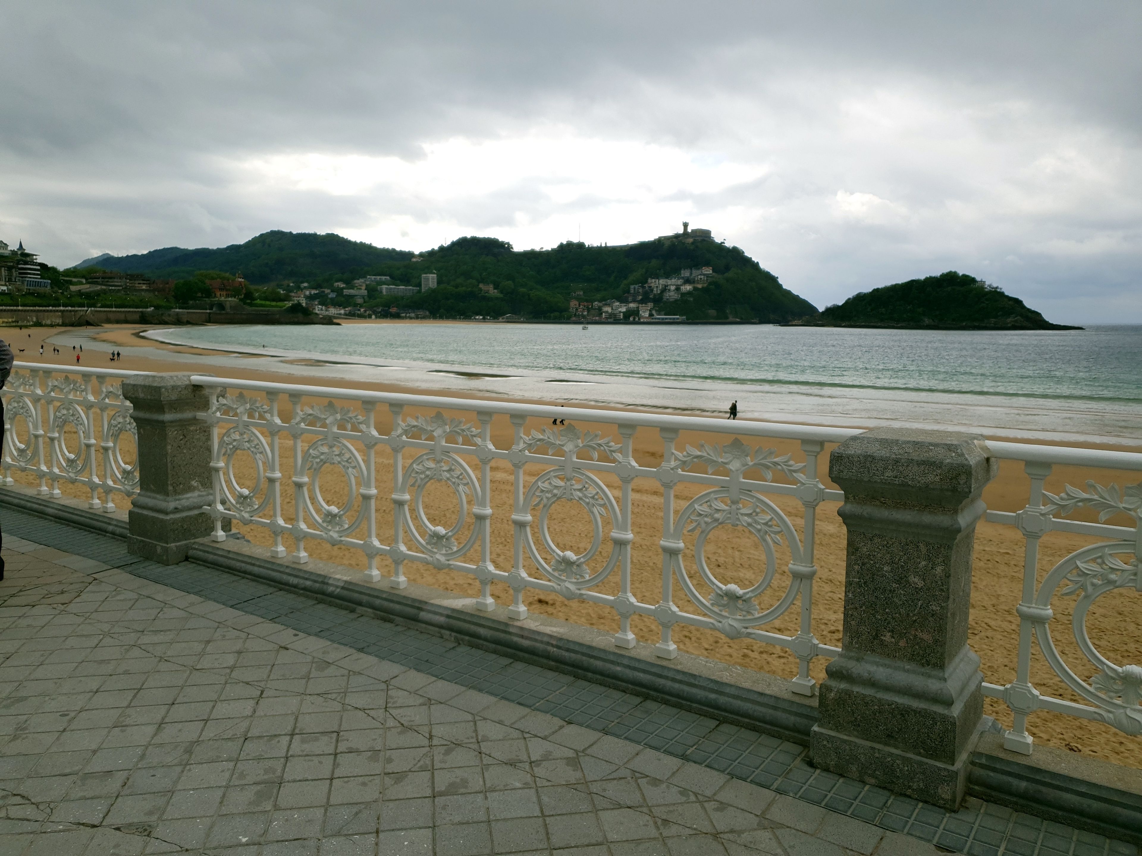 A coastal promenade with a decorative railing, a sandy beach, and a view of a hill with buildings.