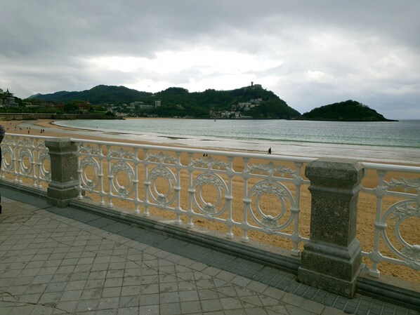 A coastal promenade with a decorative railing, a sandy beach, and a view of a hill with buildings. - Pension Beizama (Astigarraga)
