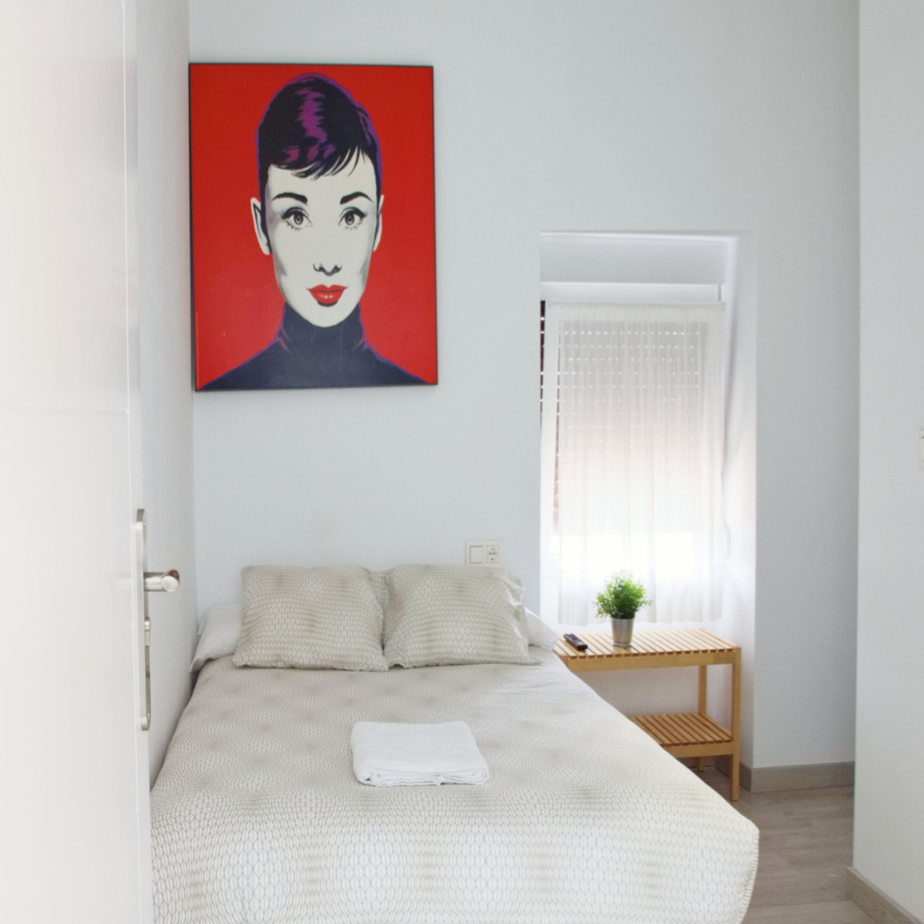 A bedroom with a bed, a window with curtains, a small table with a plant, and a painting on the wall.