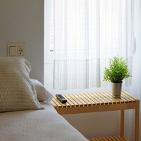 Una cama bien hecha con una colcha blanca, una mesita de noche con un control remoto y una planta en maceta en el alféizar.