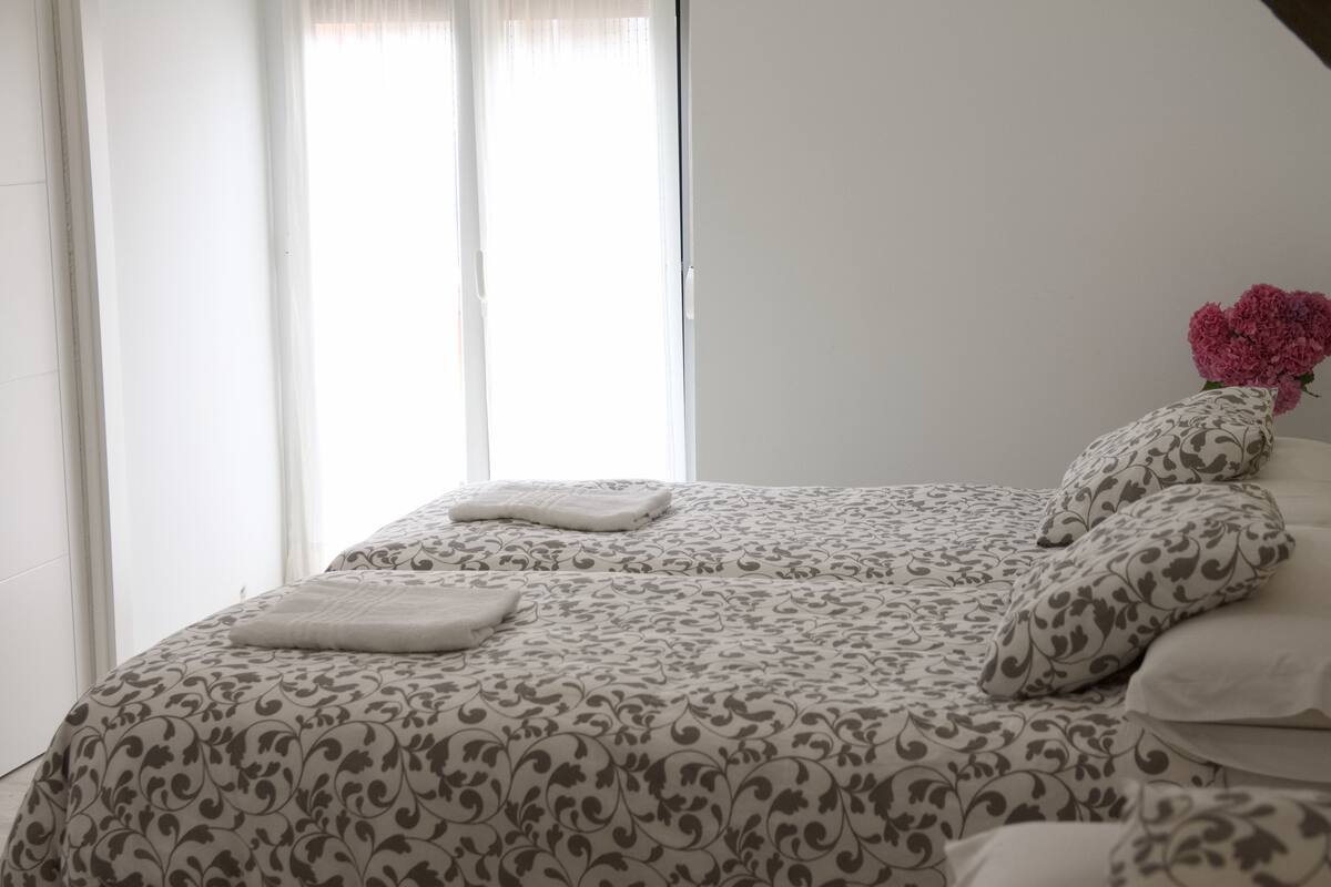 Un lit bien fait, avec une couvre-lit à motifs et des oreillers, un vase rempli de fleurs et une fenêtre ornée de rideaux légers.