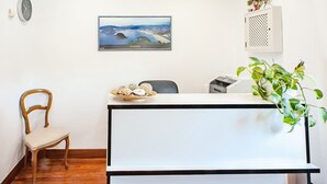 Un mostrador de recepción con una fotografía enmarcada de un paisaje, una silla de madera y una planta en maceta.