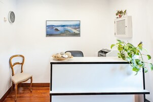 Un mostrador de recepción con una fotografía enmarcada de un paisaje, una silla de madera y una planta en maceta.