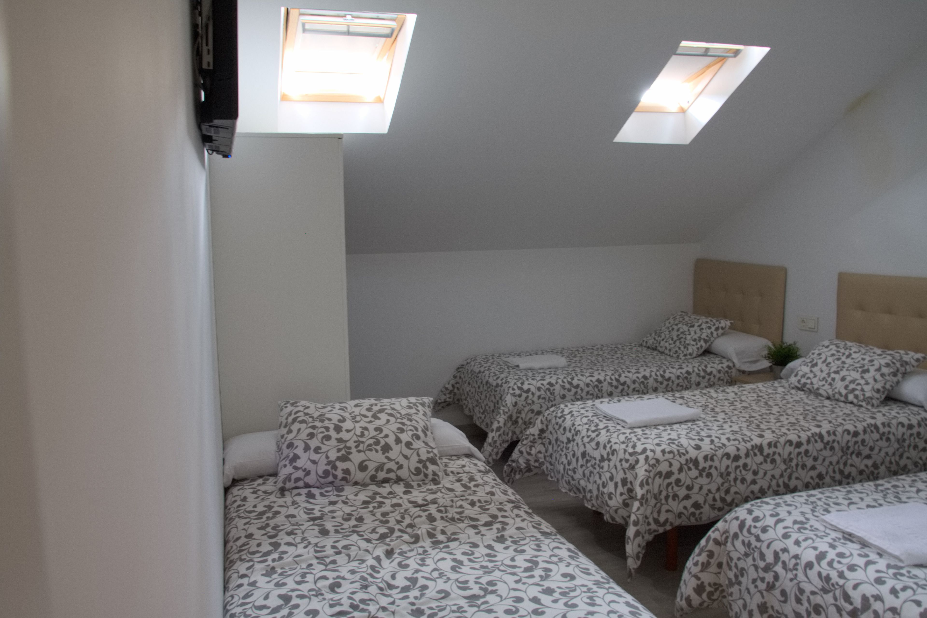 Une chambre de dortoir avec trois lits, chacun agrémenté de draps à motifs et d’un oreiller blanc. Des fenêtres de toit sont intégrées au plafond, permettant à la lumière naturelle d’entrer.