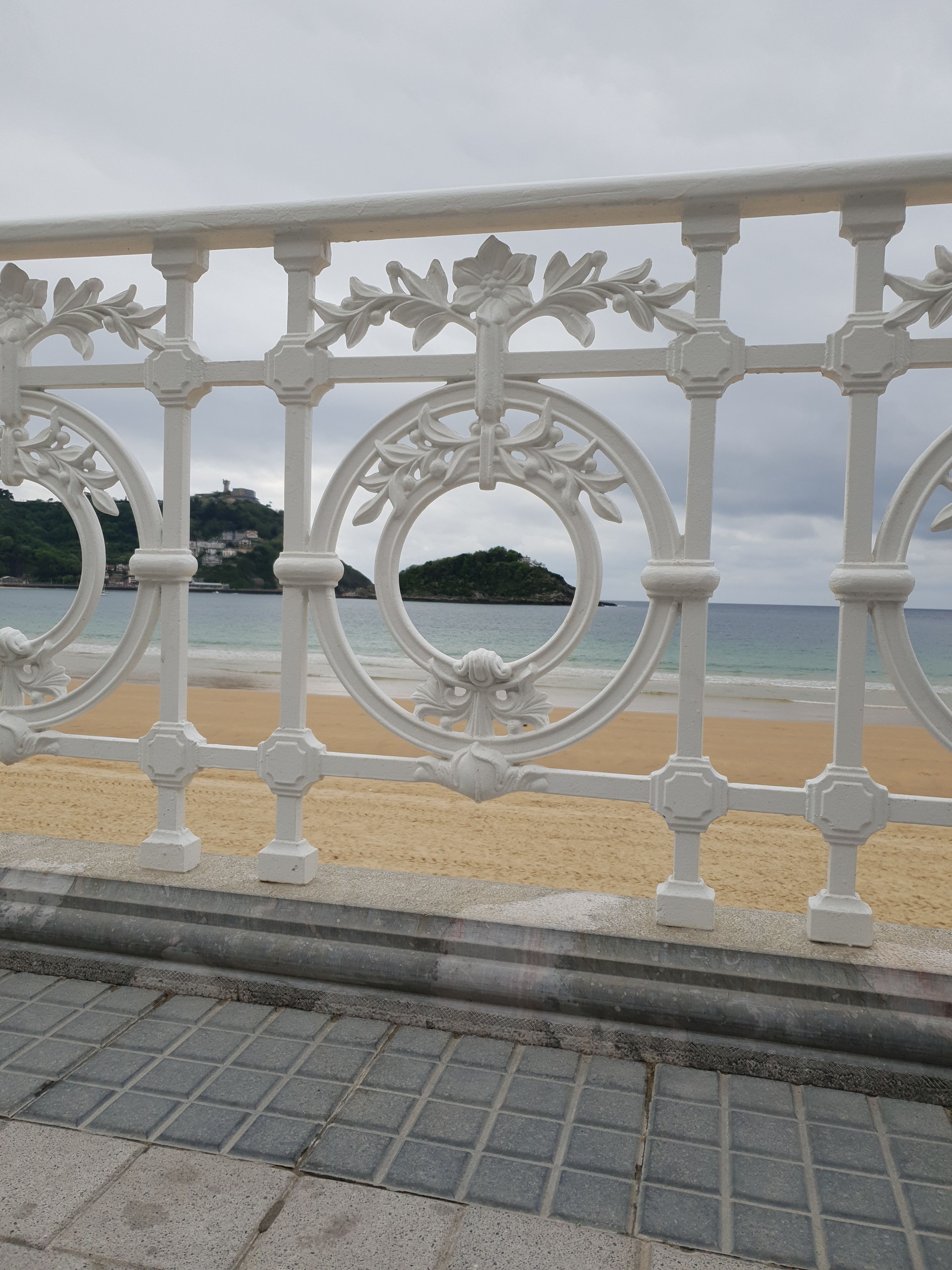 Una playa con una barandilla blanca ornamentada, una orilla arenosa y una colina distante con edificios.