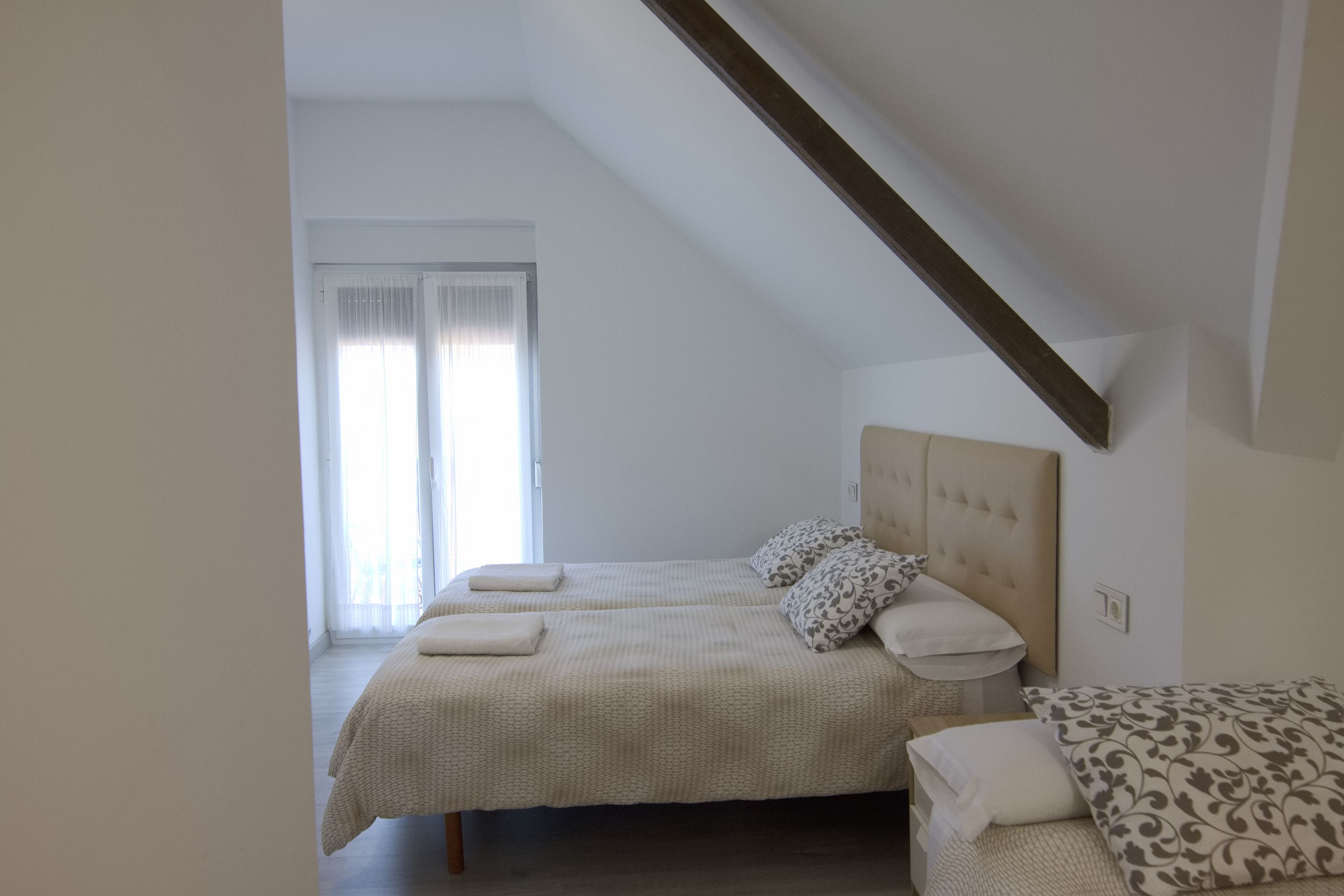 A bedroom with two beds, a sloped ceiling, and a large window with sheer curtains.