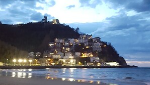 Una playa con un perro acostado sobre la arena, un litoral rocoso y un pueblo con edificios en una colina bajo un cielo nublado.