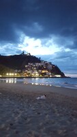 Una playa con un perro acostado sobre la arena, un litoral rocoso y un pueblo con edificios en una colina bajo un cielo nublado.