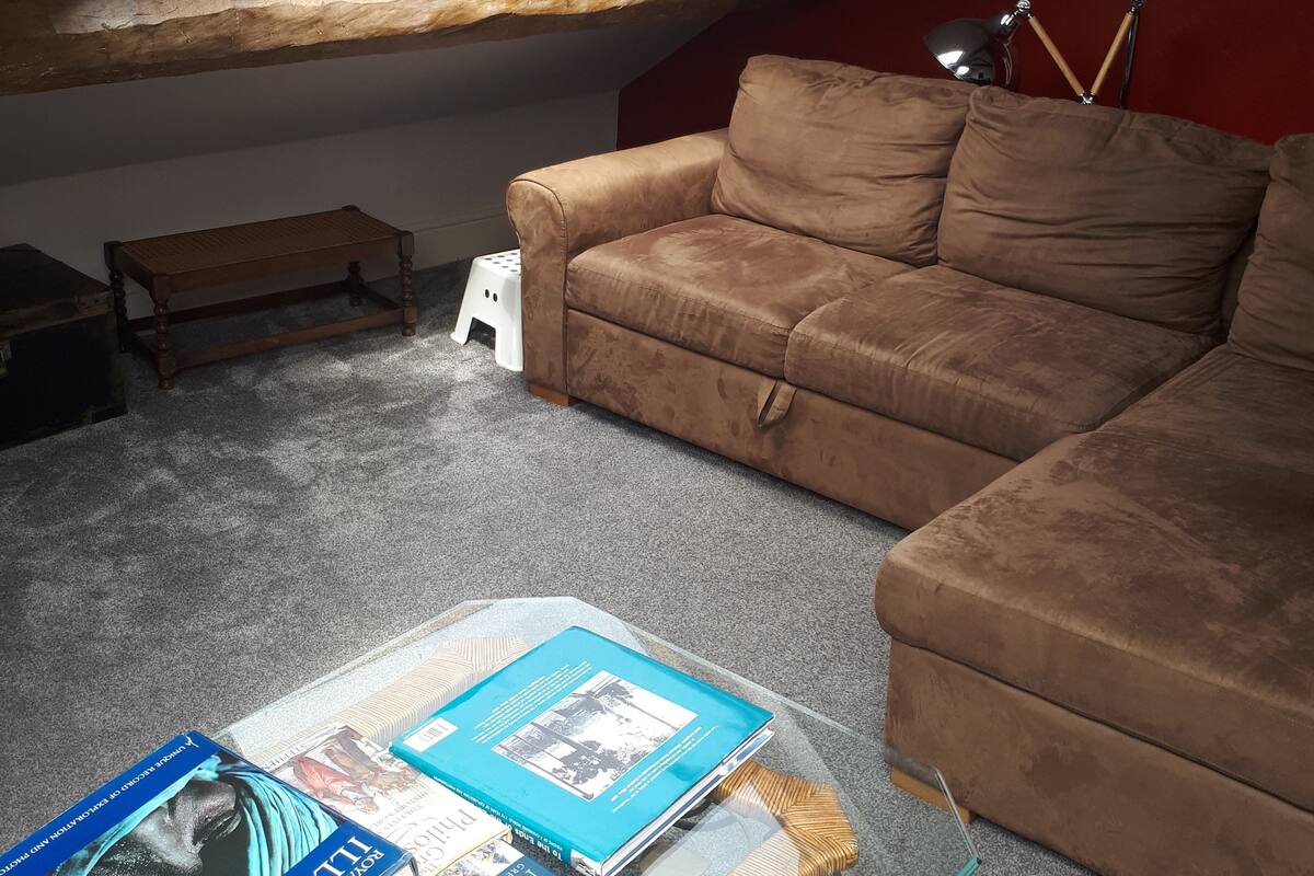 Une chambre à coucher aménagée en mezzanine, chaleureuse, avec un canapé en cuir marron, une table basse en verre sur laquelle sont posés des livres, un petit tabouret en bois et une lucarne.