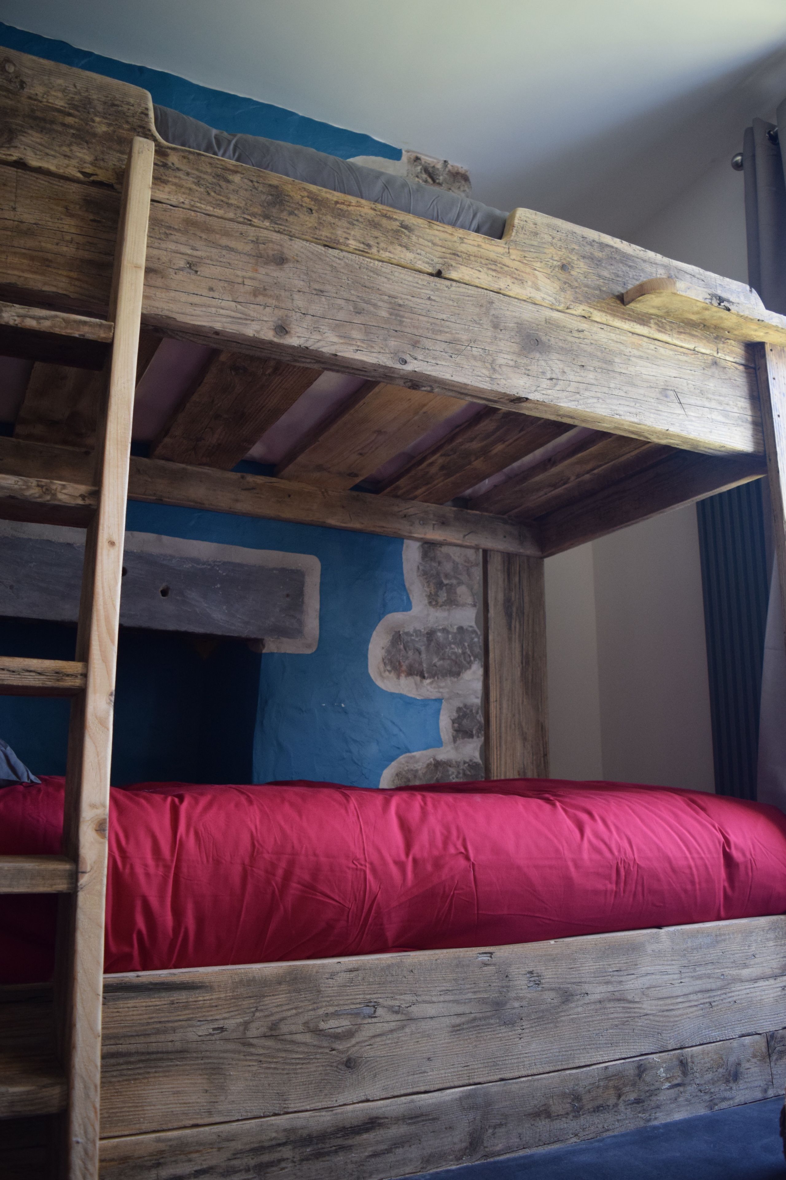 Una cama nido con colchón rojo y estructura de madera, frente a una pared azul con textura similar a la piedra.
