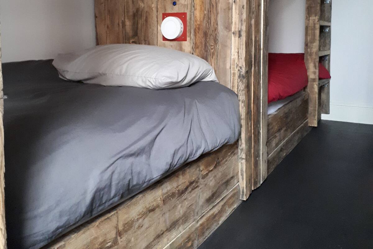 Un lit superposé avec une structure en bois, un matelas gris, des oreillers blancs et un oreiller rouge sur le lit du bas.