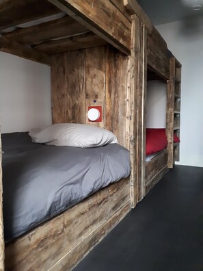 Un lit superposé avec une structure en bois, un matelas gris, des oreillers blancs et un oreiller rouge sur le lit du bas.