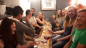 Een groep mensen zit rond een houten tafel, met glazen wijn, in een kamer met een donkere muur en een lijst met een afbeelding.