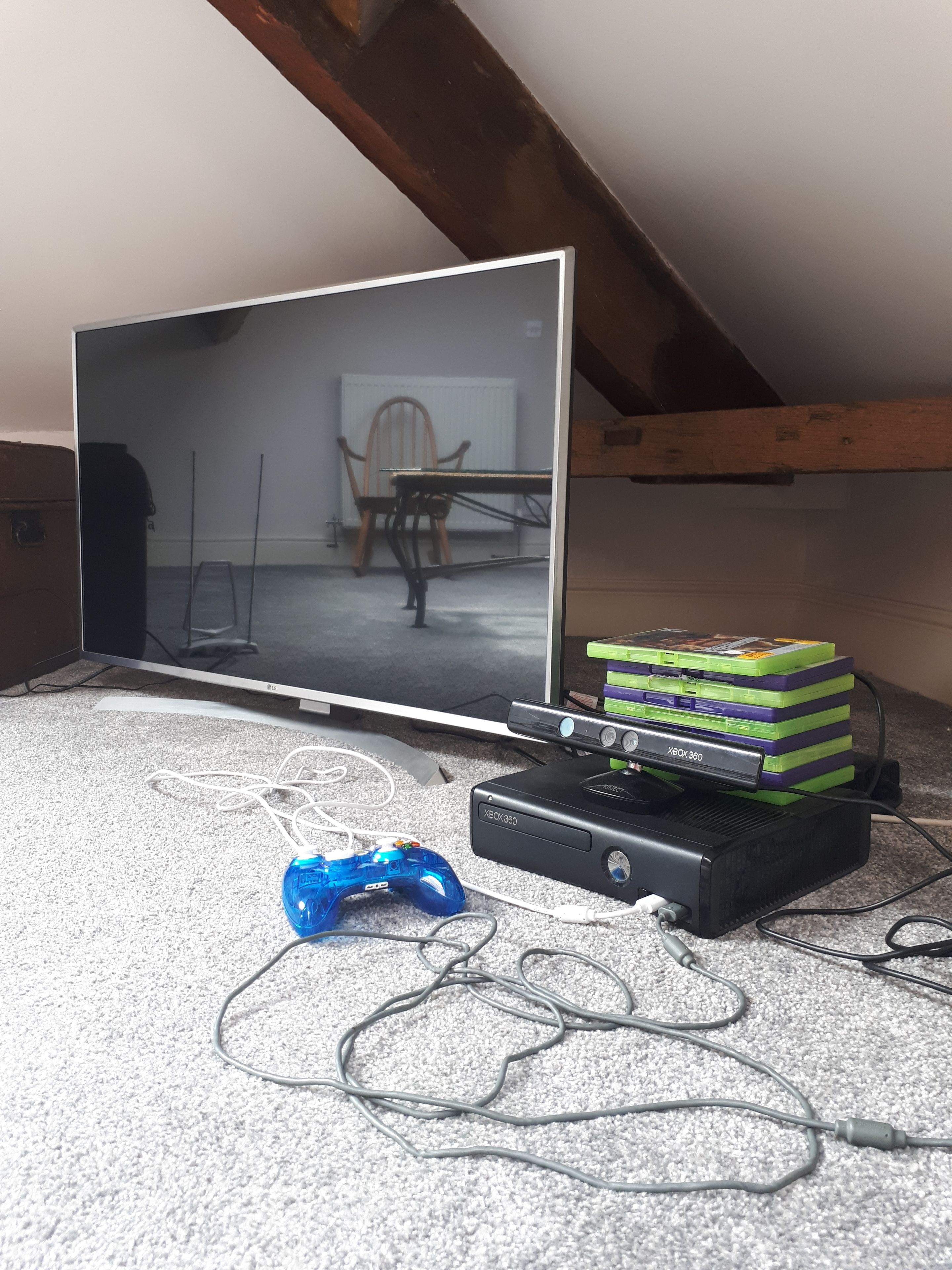 Una configuración de juegos con una consola Xbox 360, un control azul y un monitor en una habitación con techo inclinado y un radiador.