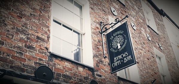 Un bâtiment en briques avec une enseigne indiquant « Byncws Caffi Bar », ornée d’un logo représentant un arbre.