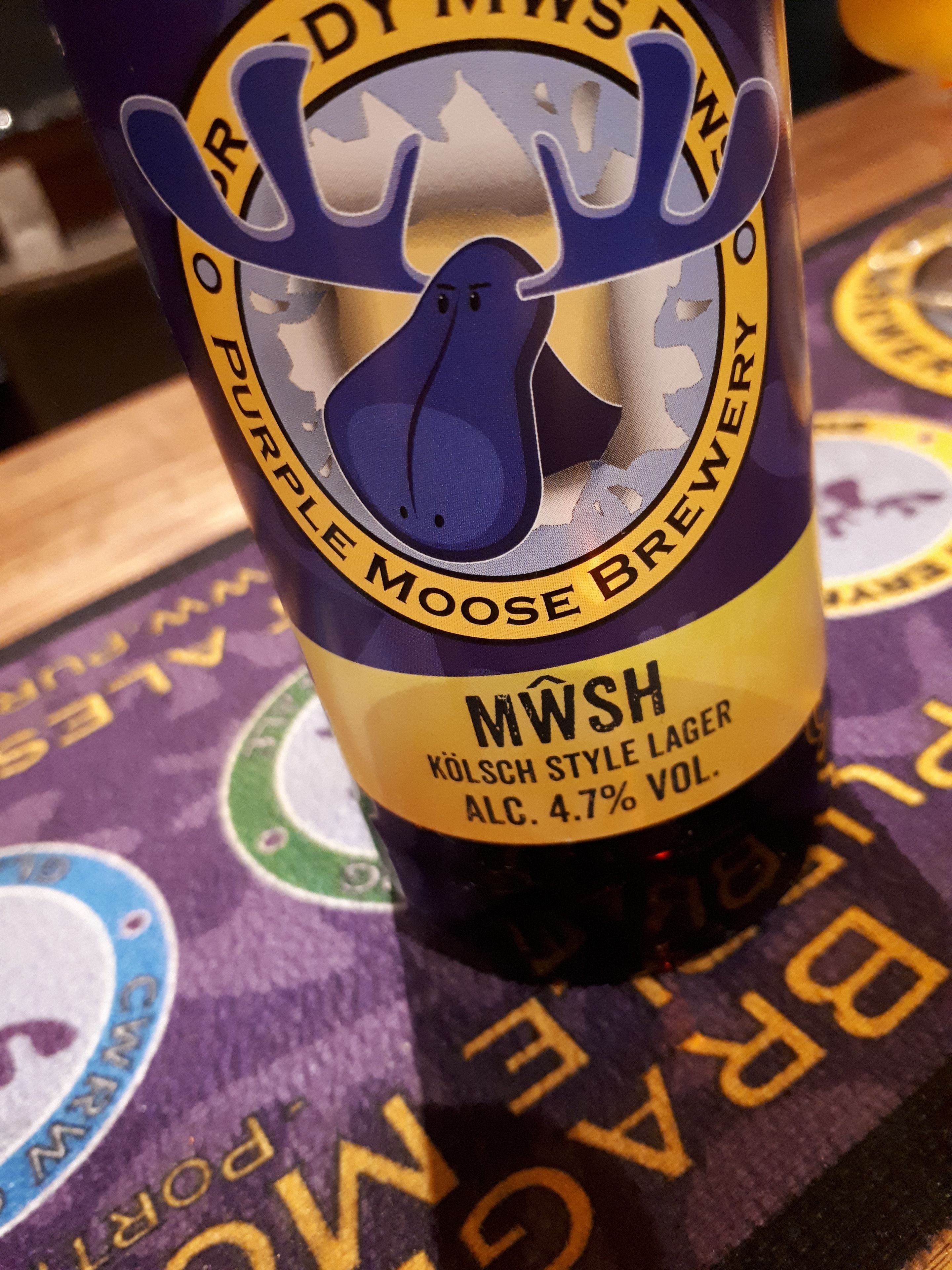 A bottle of beer with a label that reads "Purple Moose Brewery" and "MWSH Kölsch Style Lager" with an alcohol content of 4.7%.