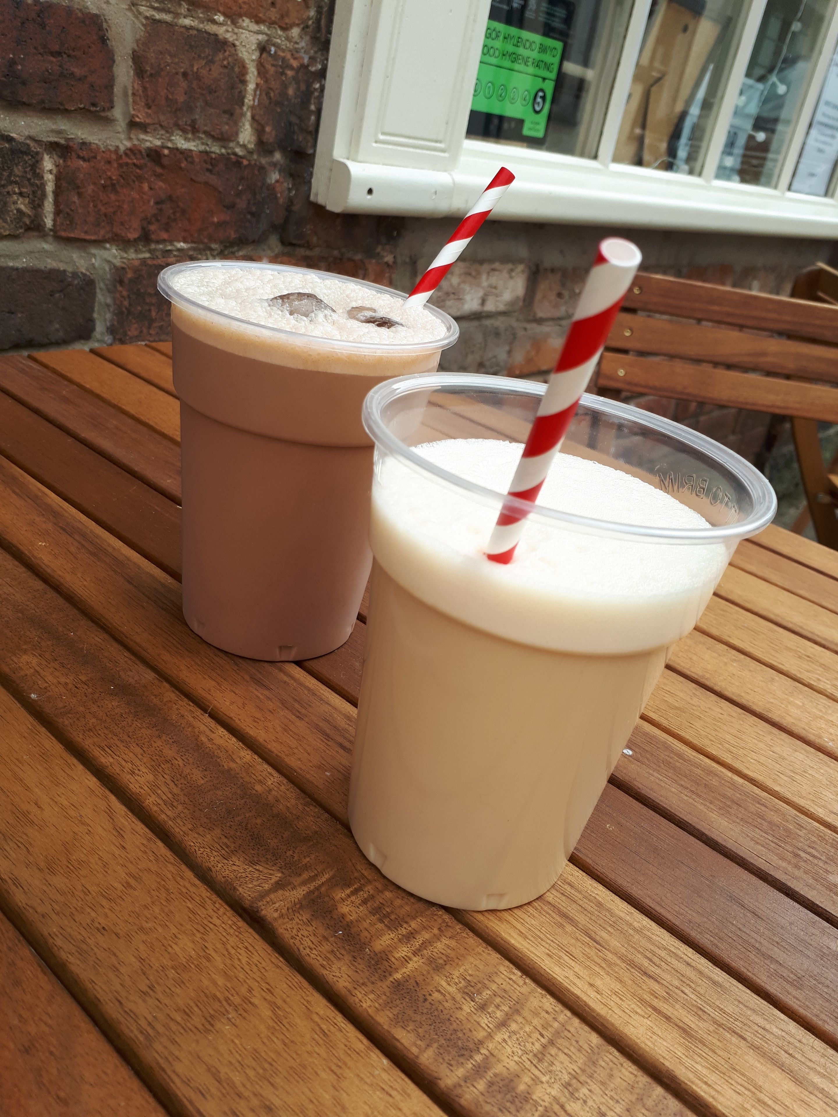 Duas bebidas em copos de plástico com canudos listrados em vermelho e branco sobre uma mesa de madeira.